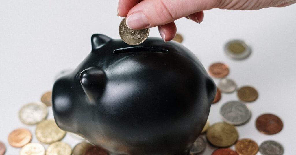 Hand inserting coin into a black piggy bank surrounded by various coins, symbolizing savings.